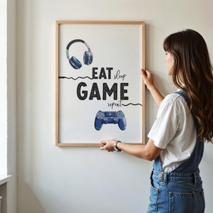 May include: A framed print with the words "EAT sleep GAME repeat" in black text. The print features illustrations of blue headphones and a game controller. The frame is a light brown colour.