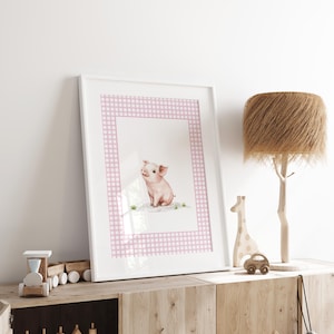 May include: A framed watercolor print featuring a charming pink piglet. The artwork is set against a white background with a pink gingham border. The print is displayed on a wooden cabinet, alongside decorative wooden toys and a lamp.