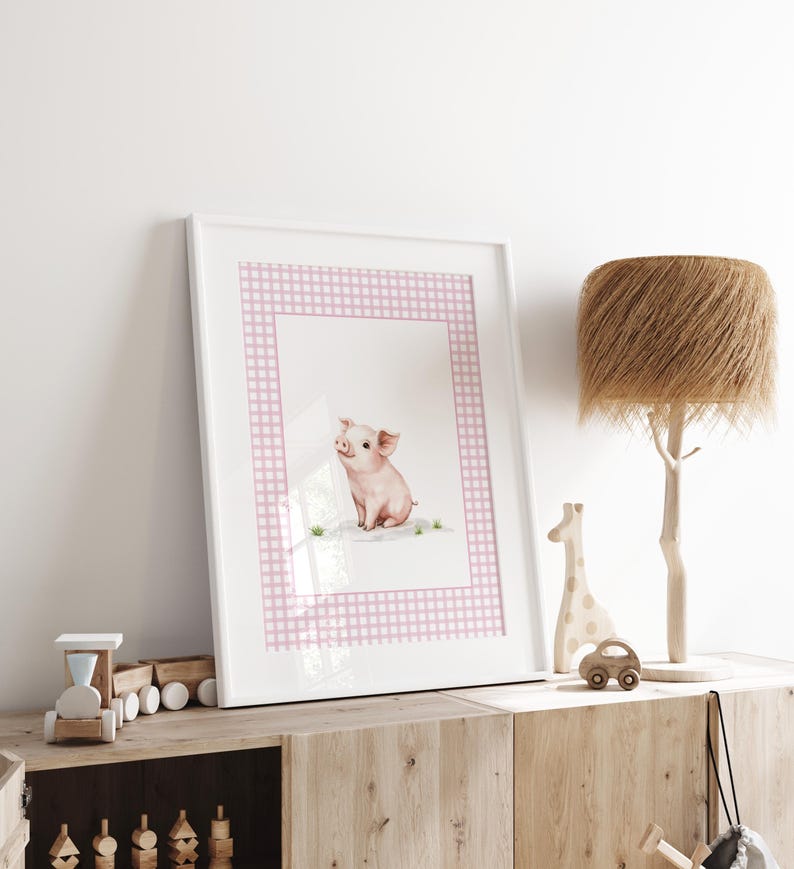 May include: A framed watercolor print featuring a charming pink piglet. The artwork is set against a white background with a pink gingham border. The print is displayed on a wooden cabinet, alongside decorative wooden toys and a lamp.