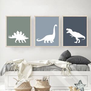 May include: Three framed prints of white dinosaur silhouettes on a light blue, dark blue, and green background. The prints are hanging on a white wall above a white bed with a white blanket and gray pillows.