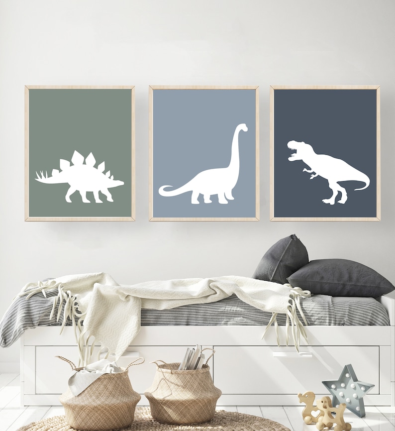 May include: Three framed prints of white dinosaur silhouettes on a light blue, dark blue, and green background. The prints are hanging on a white wall above a white bed with a white blanket and gray pillows.