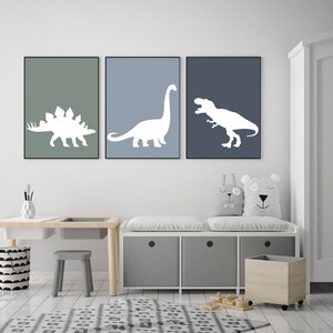 May include: Three framed prints of white dinosaur silhouettes on a blue and green background. The prints are hanging on a white wall above a white bench with storage bins and a wooden toy bin.
