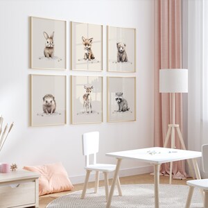 May include: A set of six framed animal prints featuring a rabbit, fox, bear, hedgehog, deer, and raccoon. The prints are in a minimalist style with a white background and gold frames. The prints are hung on a white wall with a pink curtain in the background.