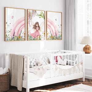 May include: Three watercolor prints featuring a fairy with pink wings standing in front of a pink rainbow with flowers and greenery. The prints are framed in wood and hung on a white wall above a white crib.