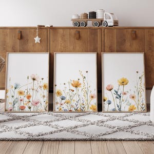 May include: Three framed watercolor prints of wildflowers in shades of yellow, orange, pink, and blue against a white background. The prints are displayed on a wooden shelf with a white rug in front.