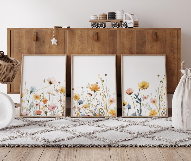 May include: Three framed watercolor prints of wildflowers in shades of yellow, orange, pink, and blue against a white background. The prints are displayed on a wooden shelf with a white rug in front.