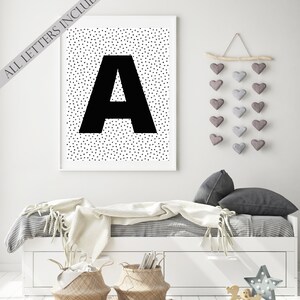 May include: A framed black and white print featuring a large black letter "A" against a background of small black triangles. The text "ALL LETTERS INCLUDED" is printed diagonally across the top left corner. The print is displayed above a white bed with decorative elements.