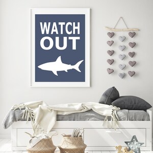 Op de afbeelding: Een wit haaien silhouet met de woorden "WATCH OUT" in witte tekst op een donkerblauwe achtergrond. De print is ingelijst in een wit frame en hangt aan een witte muur.