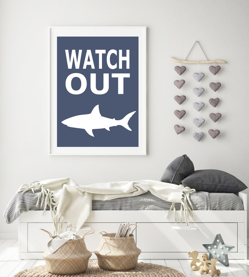 Op de afbeelding: Een wit haaien silhouet met de woorden "WATCH OUT" in witte tekst op een donkerblauwe achtergrond. De print is ingelijst in een wit frame en hangt aan een witte muur.