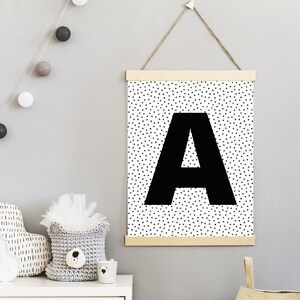 May include: A black and white wall art print featuring a large black letter "A" against a background of small black triangles. The print is suspended by a wooden frame and a natural twine cord. The theme is nursery decor.