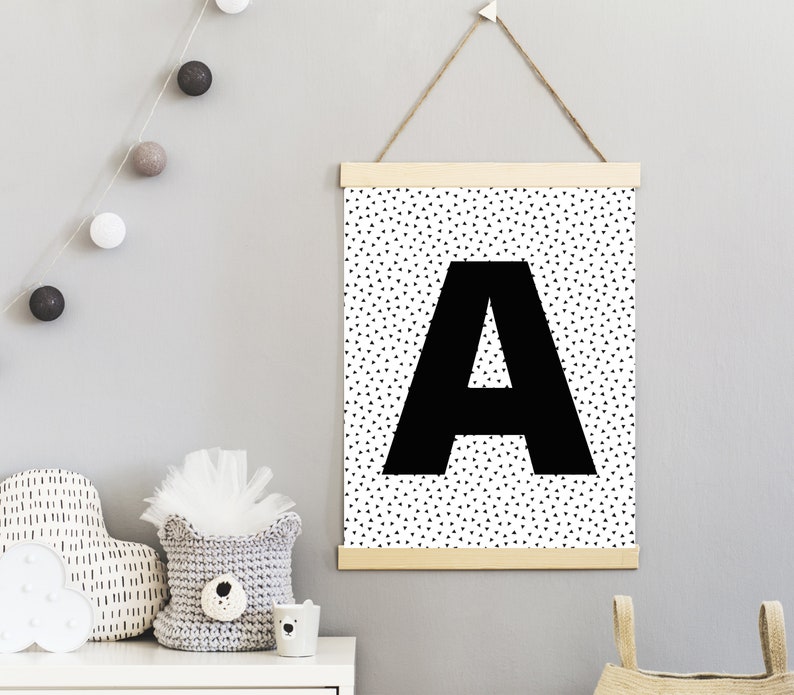 May include: A black and white wall art print featuring a large black letter "A" against a background of small black triangles. The print is suspended by a wooden frame and a natural twine cord. The theme is nursery decor.