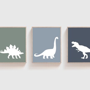 May include: Three framed prints with white dinosaur silhouettes on different colored backgrounds. The left print has a green background and a stegosaurus silhouette. The middle print has a light blue background and a brachiosaurus silhouette. The right print has a dark blue background and a tyrannosaurus rex silhouette.