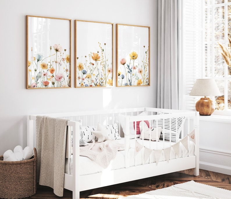 May include: Three framed watercolor paintings of floral bouquets with yellow, pink, and blue flowers on a white wall above a white crib with a white blanket and pillows.