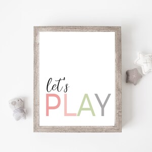 May include: A framed print with the text "let's PLAY" in pink, green, and gray letters. The print is hanging on a white wall with two gray star-shaped stuffed toys and a gray stuffed animal.