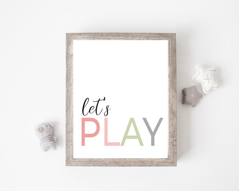 May include: A framed print with the text "let's PLAY" in pink, green, and gray letters. The print is hanging on a white wall with two gray star-shaped stuffed toys and a gray stuffed animal.