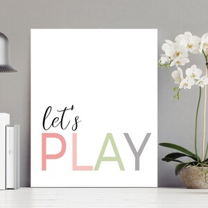 May include: A white canvas print with the text "let's PLAY" in pink, green, and gray letters. The print is hanging on a gray wall with a silver pendant light above it and a potted white orchid plant to the right.