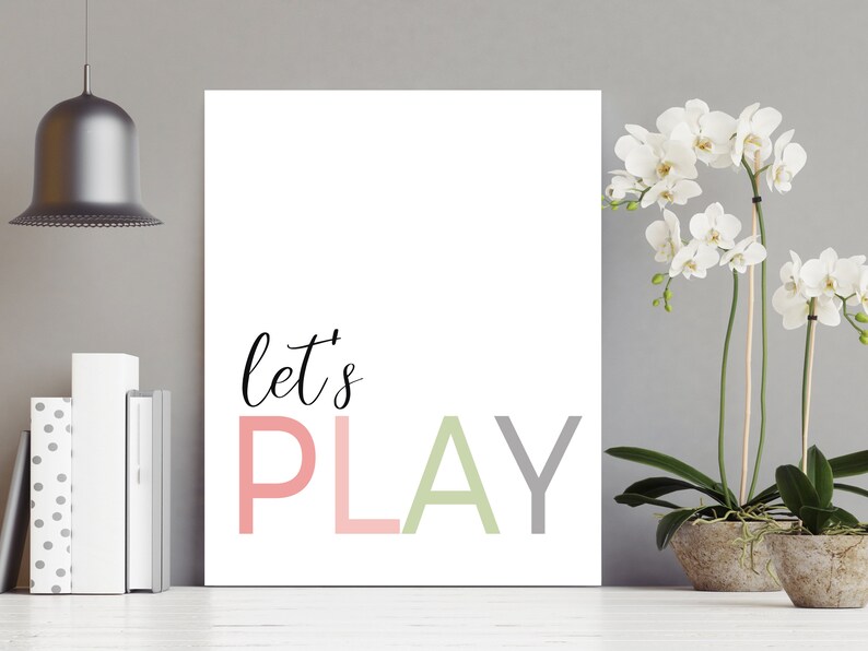 May include: A white canvas print with the text "let's PLAY" in pink, green, and gray letters. The print is hanging on a gray wall with a silver pendant light above it and a potted white orchid plant to the right.