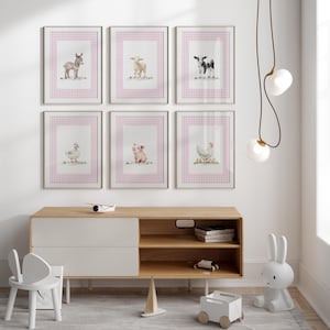 May include: Six framed watercolor prints of farm animals: a donkey, sheep, cow, duck, pig, and chicken. Each print features the animal on a white background with a pink gingham border. The prints are displayed above a wooden cabinet.