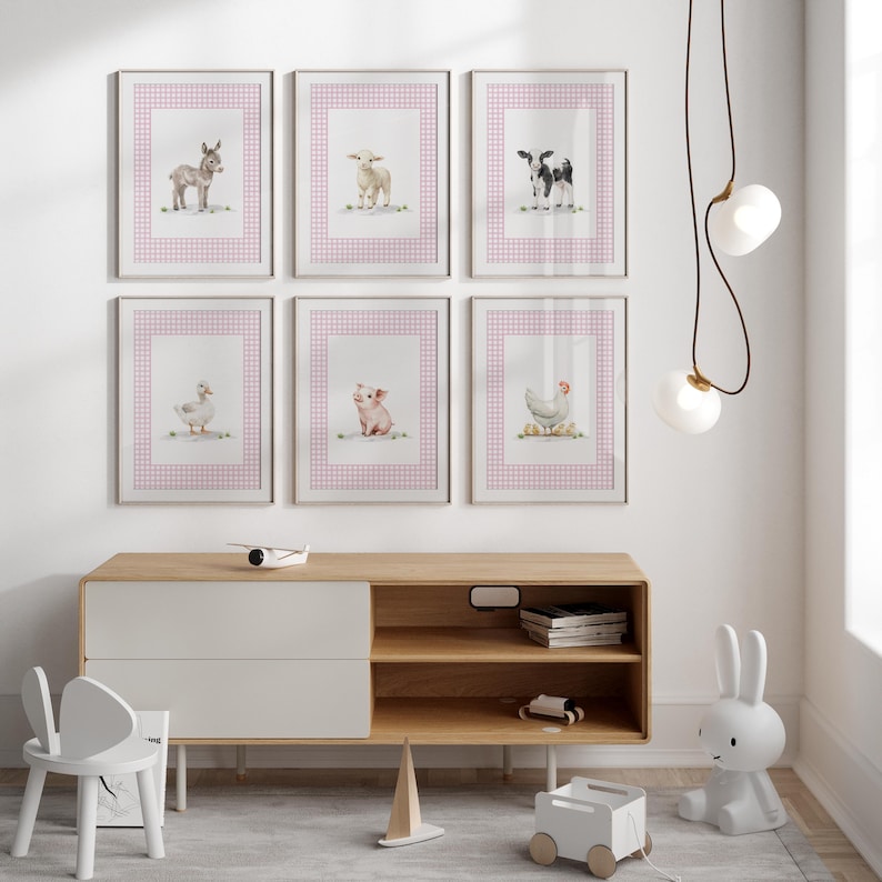 May include: Six framed watercolor prints of farm animals: a donkey, sheep, cow, duck, pig, and chicken. Each print features the animal on a white background with a pink gingham border. The prints are displayed above a wooden cabinet.