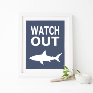 Op de afbeelding: Een wit ingelijst print met een blauwe achtergrond en witte tekst die "WATCH OUT" luidt. Onder de tekst bevindt zich een wit silhouet van een haai.