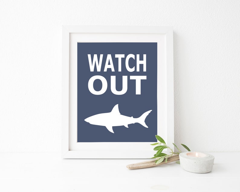 Op de afbeelding: Een wit ingelijst print met een blauwe achtergrond en witte tekst die "WATCH OUT" luidt. Onder de tekst bevindt zich een wit silhouet van een haai.