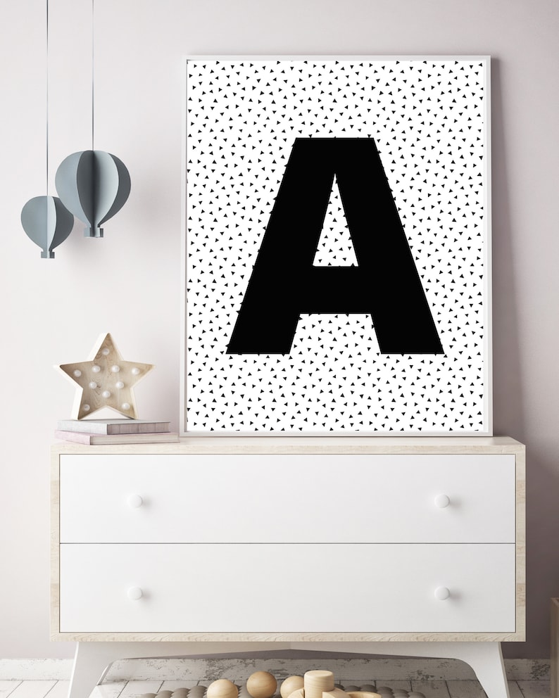 May include: A framed black and white print featuring a large black letter "A" against a background of small black triangles. The artwork is displayed on a white dresser with decorative items, including a star-shaped light and paper hot air balloons.