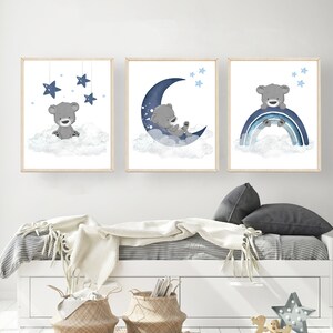 Op de afbeelding: Drie ingelijste prints met een grijze beer op een witte wolk met blauwe sterren, een grijze beer op een blauwe halve maan met witte sterren en een grijze beer op een blauwe regenboog met witte sterren.