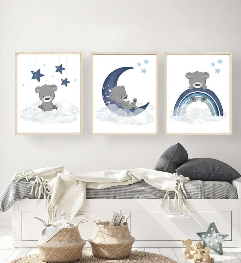 Op de afbeelding: Drie ingelijste prints met een grijze beer op een witte wolk met blauwe sterren, een grijze beer op een blauwe halve maan met witte sterren en een grijze beer op een blauwe regenboog met witte sterren.