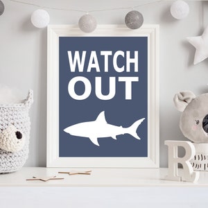 Op de afbeelding: Een wit ingelijst afdruk met een blauwe achtergrond en witte tekst die "WATCH OUT" zegt. Een wit silhouet van een haai bevindt zich in het midden van de afdruk.