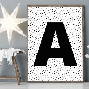 May include: A framed art print featuring a large black letter "A" against a white background covered in small black triangles. The artwork is displayed in a room with a wooden stool, a star-shaped light fixture, and a vase with white flowers.