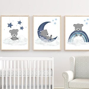 Op de afbeelding: Drie ingelijste prints met een blauw en grijs kleurenschema met een schattige grijze beer op een wolk met sterren, een grijze beer op een halve maan met sterren en een grijze beer op een regenboog met sterren.