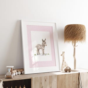 May include: A framed watercolor print of a baby donkey. The artwork features a light gray donkey standing on a grassy area, set against a pink gingham border. The print is displayed in a white frame and rests on a wooden cabinet.