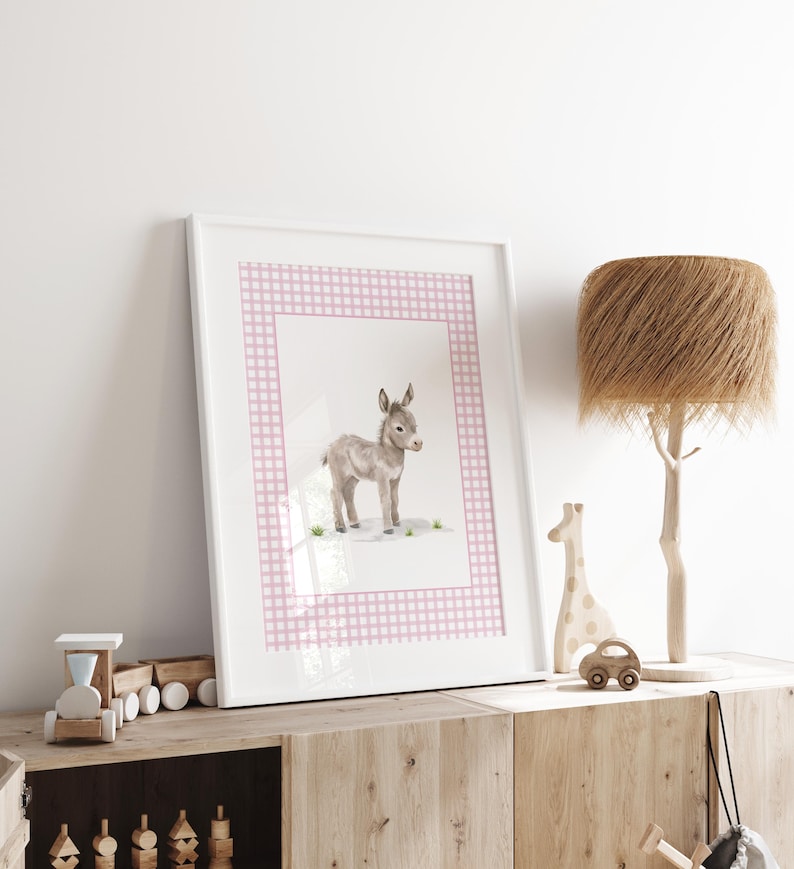 May include: A framed watercolor print of a baby donkey. The artwork features a light gray donkey standing on a grassy area, set against a pink gingham border. The print is displayed in a white frame and rests on a wooden cabinet.
