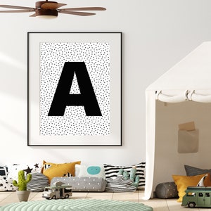 May include: A framed black and white print featuring a large black letter "A" against a background of small black triangles. The print hangs above a collection of colorful pillows, a green rug, and toy vehicles, creating a playful nursery or playroom setting.