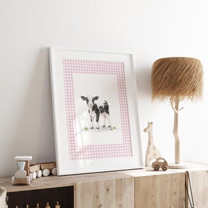 May include: A framed watercolor print of a black and white calf, set against a pink gingham border. The artwork is displayed on a wooden cabinet, alongside a wooden train, a giraffe, and a car, with a decorative lamp.