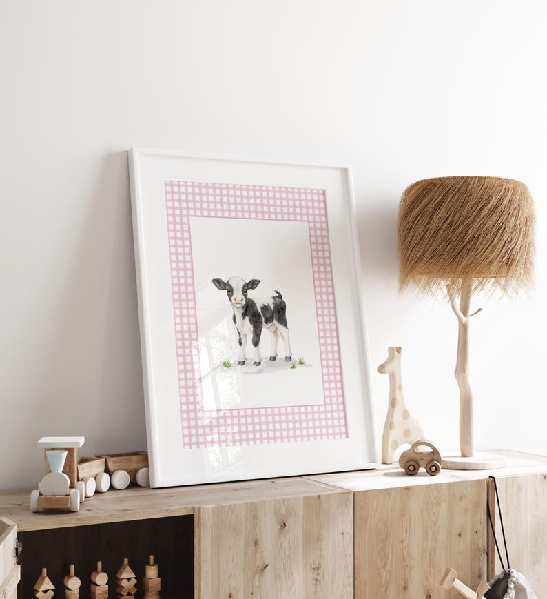 May include: A framed watercolor print of a black and white calf, set against a pink gingham border. The artwork is displayed on a wooden cabinet, alongside a wooden train, a giraffe, and a car, with a decorative lamp.