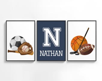 May include: Three framed watercolor prints featuring sports equipment. One shows a soccer ball, baseball glove, and bat. Another displays a basketball, hockey stick, and football. The center print has the name "NATHAN" and the letter "N".