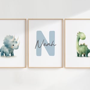 Może przedstawiać: Trzy oprawione wydruki akwarelowe z dinozaurami. Jeden przedstawia niebieskiego triceratopsa, drugi literę "N" z imieniem "Noah", a trzeci zielonego Brontozaura. Wszystkie na białym tle.
