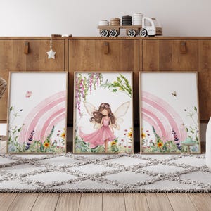 May include: Three watercolor prints featuring a fairy with wings and a pink dress standing in front of a pink rainbow with flowers and greenery. The prints are framed in white and hung on a wooden wall.