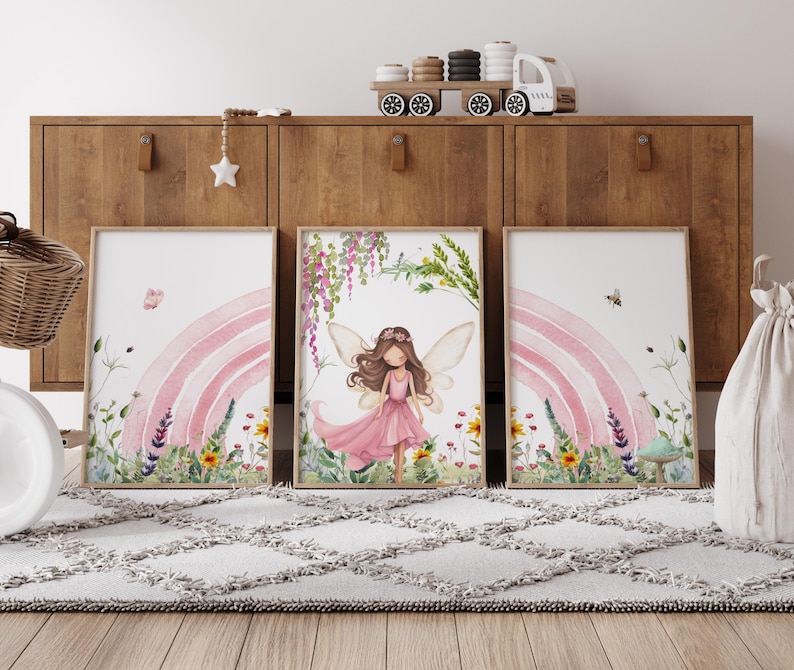 May include: Three watercolor prints featuring a fairy with wings and a pink dress standing in front of a pink rainbow with flowers and greenery. The prints are framed in white and hung on a wooden wall.