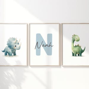 May include: Three framed art prints featuring watercolor dinosaurs and the name "Noah". The first print shows a blue triceratops, the second a blue letter "N" with the name "Noah", and the third a green dinosaur.