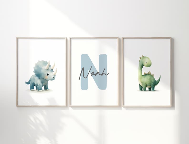Może przedstawiać: Trzy oprawione wydruki artystyczne z akwarelowymi ilustracjami dinozaur&oacute;w i imieniem "Noah". Pierwszy wydruk przedstawia niebieskiego triceratopsa, drugi niebieską literę "N" z imieniem "Noah", a trzeci zielonego dinozaura.
