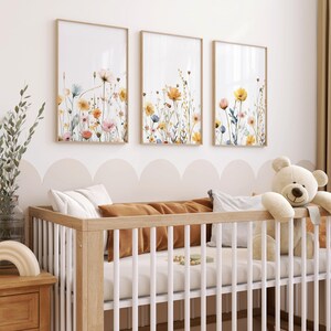 May include: A white crib with a brown wooden frame and a white mattress. The crib is decorated with three framed prints of watercolor floral arrangements in shades of yellow, orange, and pink. A white teddy bear is sitting on the edge of the crib.