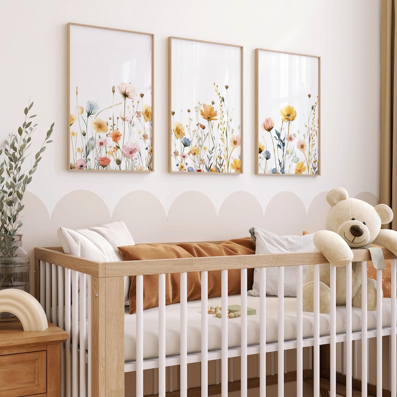 May include: A white crib with a brown wooden frame and a white mattress. The crib is decorated with three framed prints of watercolor floral arrangements in shades of yellow, orange, and pink. A white teddy bear is sitting on the edge of the crib.