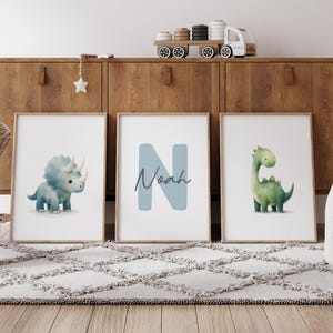 Może przedstawiać: Trzy oprawione grafiki z dinozaurami. Jedna przedstawia niebieskiego triceratopsa, druga ma imię "Noah" z niebieską literą "N", a trzecia przedstawia zielonego Brontozaura. Grafiki są wystawione na drewnianej szafce.