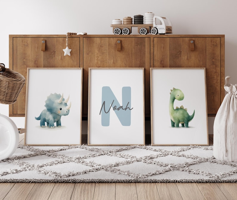 Może przedstawiać: Trzy oprawione grafiki z dinozaurami. Jedna przedstawia niebieskiego triceratopsa, druga ma imię "Noah" z niebieską literą "N", a trzecia przedstawia zielonego Brontozaura. Grafiki są wystawione na drewnianej szafce.