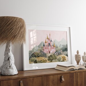 May include: A framed watercolor print of a pink castle with gold and blue turrets, set against a backdrop of green hills and a pink sky. The artwork is displayed on a wooden cabinet, next to a lamp and an open book.