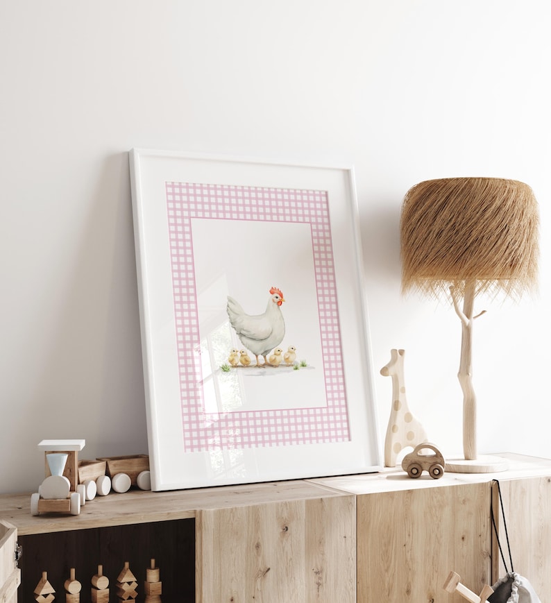 May include: A framed print featuring a watercolor illustration of a white hen with a red comb and four yellow chicks. The artwork is set against a pink and white gingham border. The print is displayed on a wooden cabinet with decorative items.