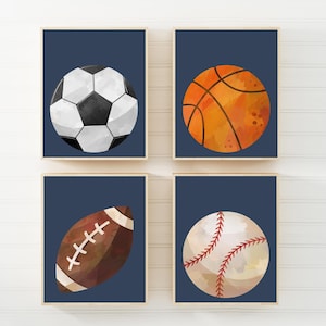 May include: Four framed prints featuring sports balls. A football with black and white panels, an orange basketball, a brown rugby ball, and a baseball with red stitching, all on a navy blue background.