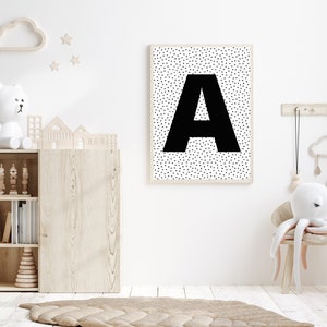 May include: A framed print featuring a large black letter "A" against a white background speckled with small black triangles. The print hangs on a white wall in a child's room, alongside wooden toys and a rug.